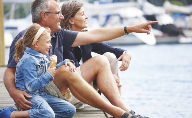 Happy Grandparents sitting with their granddaughter on a pier, pointing out at the boats - copyspace